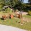 Parco giochi all'aperto 'Trixie' con strutture di arrampicata in legno e una sabbiera circolare in un parco verde.
