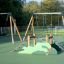 Double Safety Seat double swing in un parco giochi con struttura da arrampicata sullo sfondo.