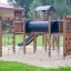 Attrezzatura da gioco in legno Romping Cottage con reti da arrampicata, scale e scivolo in un parco.