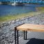 Moderno Thor long bench in legno con gambe in metallo lungo un sentiero sul fiume con un ponte blu sullo sfondo