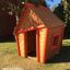 Casetta in legno per bambini - Crooked House, marrone con rifiniture rosse, in ambiente esterno.