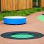 Round trampoline (100 cm) integrato nella superficie del parco giochi con tappeto elastico verde e bordo nero.
