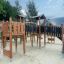 Struttura moderna in legno per parco giochi con scivolo ed elementi per arrampicarsi, circondata da sabbia e alberi.