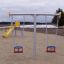 Baby Double Swing in un parco giochi con superficie in sabbia e scivolo giallo sullo sfondo.