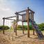 Kampinos wooden playhouse con scivoli e rete da arrampicata in un parco giochi sabbioso.