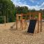 Attrezzatura da parco giochi in legno Alstad con scivolo, parete da arrampicata e altalene in un contesto di parco.