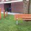 Parco giochi Triple Somersault Nature vicino a un edificio in mattoni con panchina in legno e attrezzature da palestra all'aperto.
