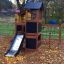 Casetta gioco in legno con scivolo in un parco giochi circondato da foglie autunnali.