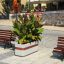 Fioriera Geneva IV con rigoglioso fogliame verde e fiori arancioni su una piazza pubblica pavimentata tra elementi in legno