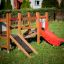 Mini play set da gioco in legno con uno scivolo rosso e una rampa da arrampicata su un prato verde.