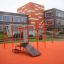 Stazione fitness all'aperto New Age Fit MEDIUM con strutture arancioni davanti a un edificio moderno.