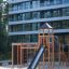 Struttura da parco giochi in legno Explorer castle with slide su area sabbiosa davanti a un edificio moderno