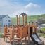 Rubik Modern Nature wooden playhouse with slide su un parco giochi sabbioso