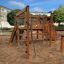 Kiosk Modern Nature wooden playhouse in un parco giochi sabbioso circondato da edifici e alberi.
