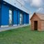 Struttura in legno Nature Playhouse su un prato erboso davanti a un moderno edificio blu.