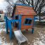 Casetta da gioco in legno Olle's Playhouse con dettagli blu e scivolo in un parco innevato.